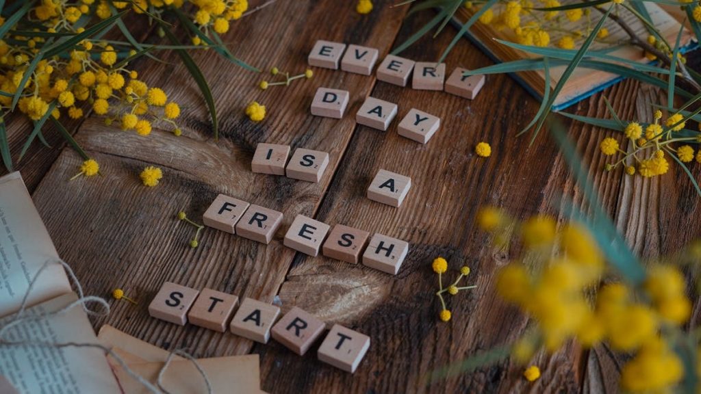 Wooden tiles spell 'Every day is a fresh start' surrounded by yellow flowers on rustic table.