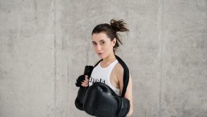 Confident young woman posing with boxing gloves against a concrete wall, showcasing style and sportiness.