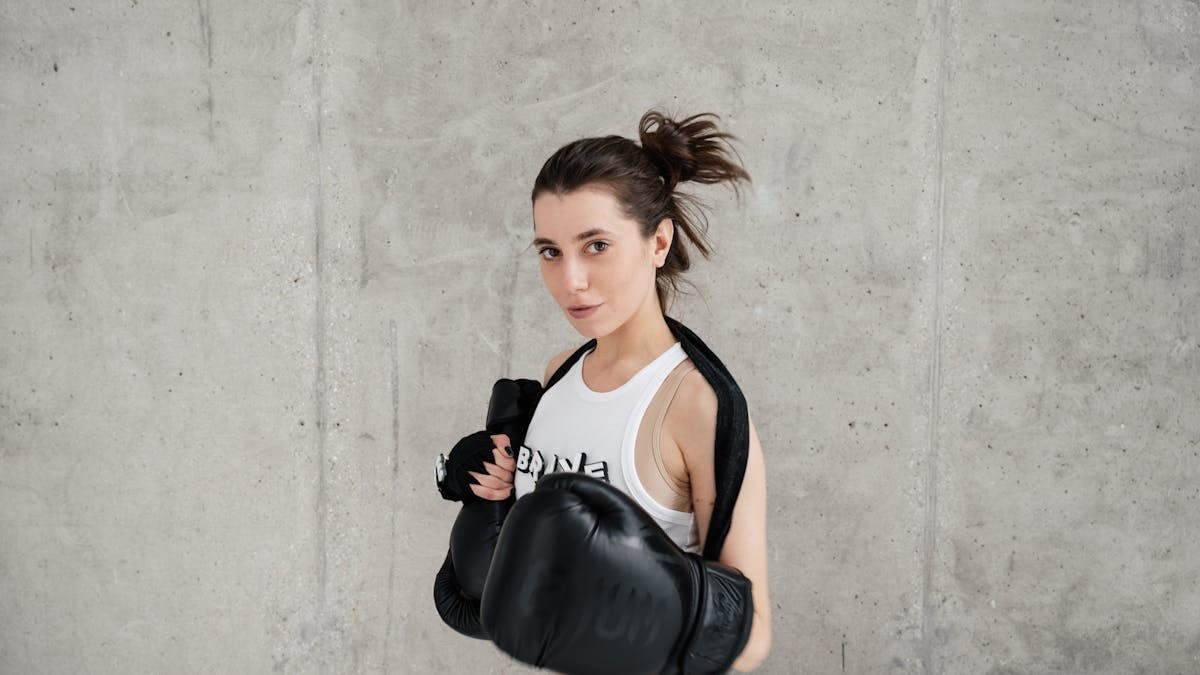 Confident young woman posing with boxing gloves against a concrete wall, showcasing style and sportiness.